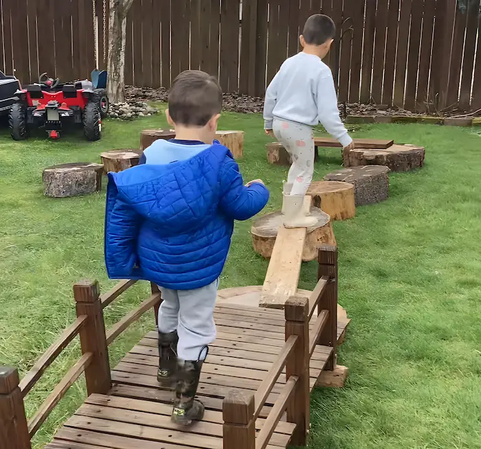 A child carefully balancing while walking on wooden logs in the outdoor play area, improving gross motor skills.