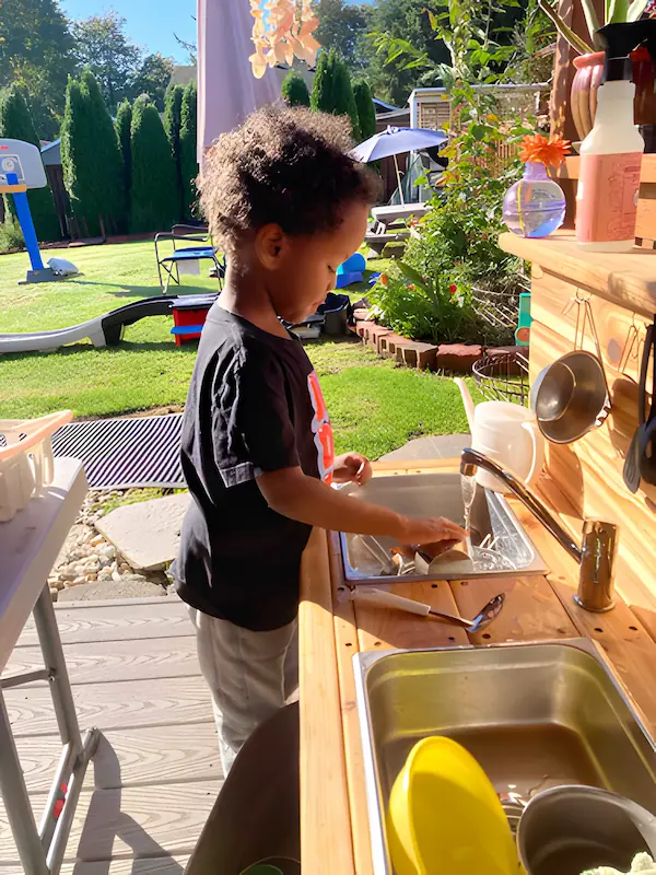 A young toddler independently washing dishes in a child-sized sink, developing practical life skills.
